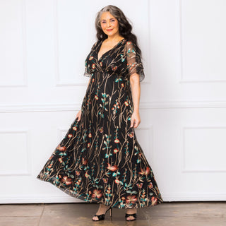 The plus size model is wearing the Embroidered Elegance Evening Gown by Kiyonna.  It is a full length black dress with colorful floral and butterfly embroidery throughout.