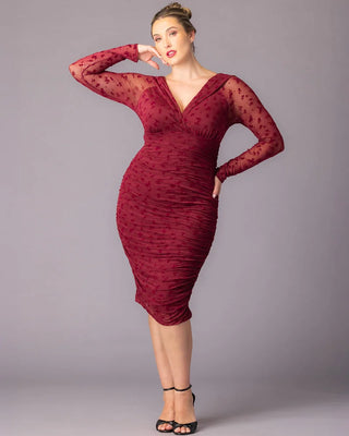 Betsey Ruched Dress in Burgundy Flocked Florals  A plus-size model poses in a fitted burgundy cocktail dress with long sheer mesh sleeves and embroidered floral details. The dress features a ruched silhouette and a deep V-neckline. She stands against a gray studio background with one arm bent near her head.