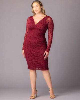 Betsey Ruched Dress in Burgundy Flocked Florals A plus-size model stands in a burgundy ruched cocktail dress with long sheer embroidered sleeves and a V-neck bodice. She wears gold heels and poses against a gray backdrop with her arms relaxed at her sides.