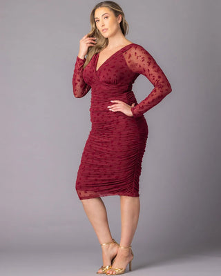 Betsey Ruched Dress in Burgundy Flocked Florals A plus-size model poses in a burgundy fitted cocktail dress featuring floral mesh long sleeves, a V-neckline, and allover ruching. She stands in a three-quarter angle with one hand on her hip against a gray studio background.