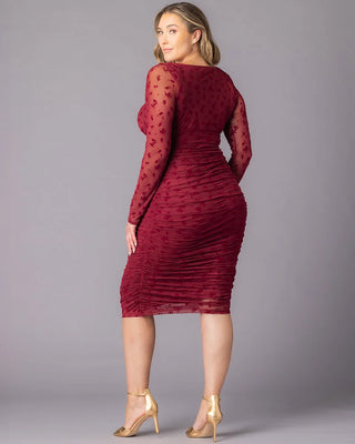 Betsey Ruched Dress in Burgundy Flocked Florals Back view of a plus-size model wearing a burgundy ruched cocktail dress with sheer floral long sleeves. She looks back over her shoulder while standing against a gray studio backdrop.