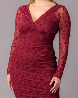 Betsey Ruched Dress in Burgundy Flocked Florals Close-up of a plus-size model wearing a burgundy ruched cocktail dress. The image showcases the deep V-neckline, gathered bodice, and long sheer mesh sleeves with flocked floral design.